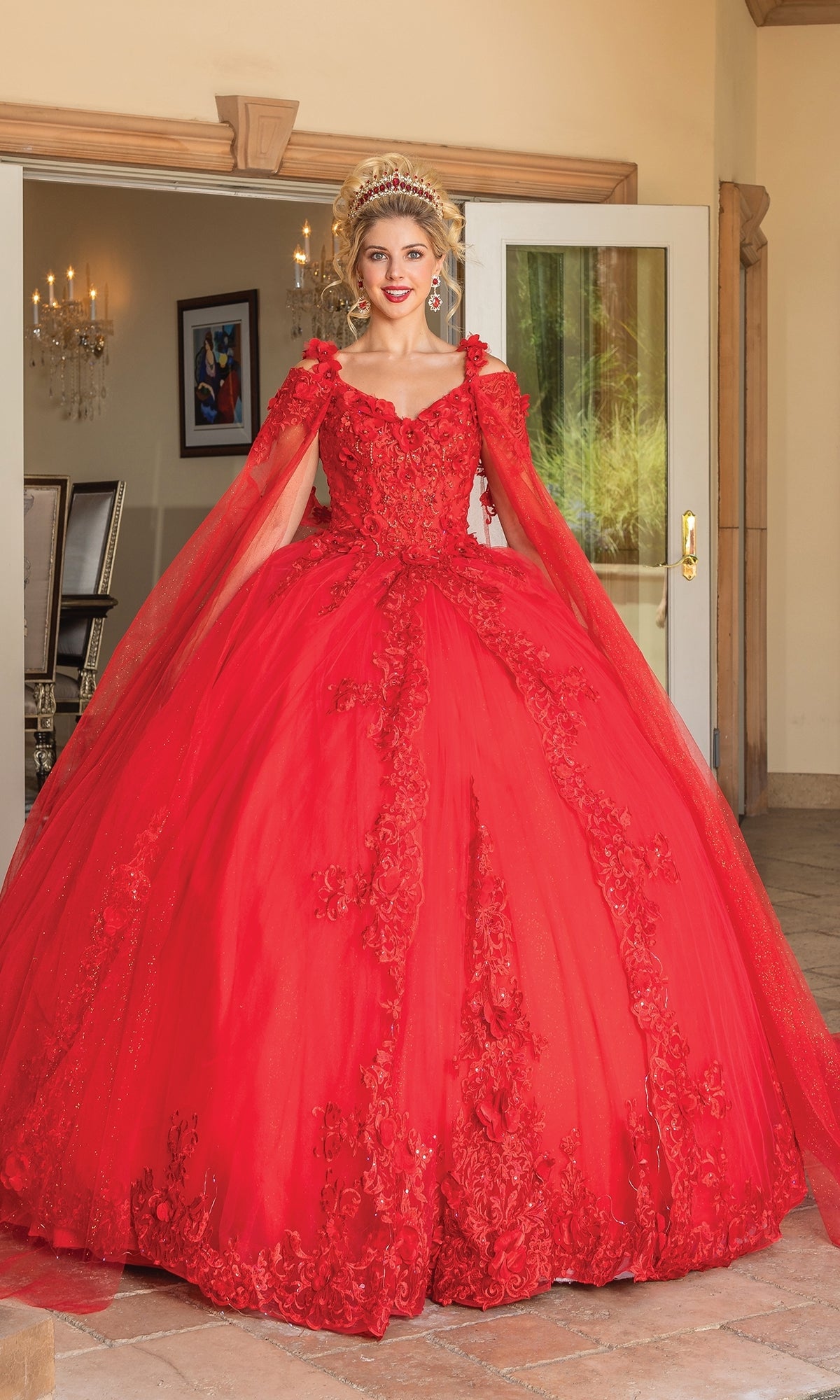 A woman stands indoors wearing the Quinceanera Dress 1678 by Dancing Queen, a bright red ball gown with floral details, matching cape, removable draped sleeves, plus a tiara and earrings.